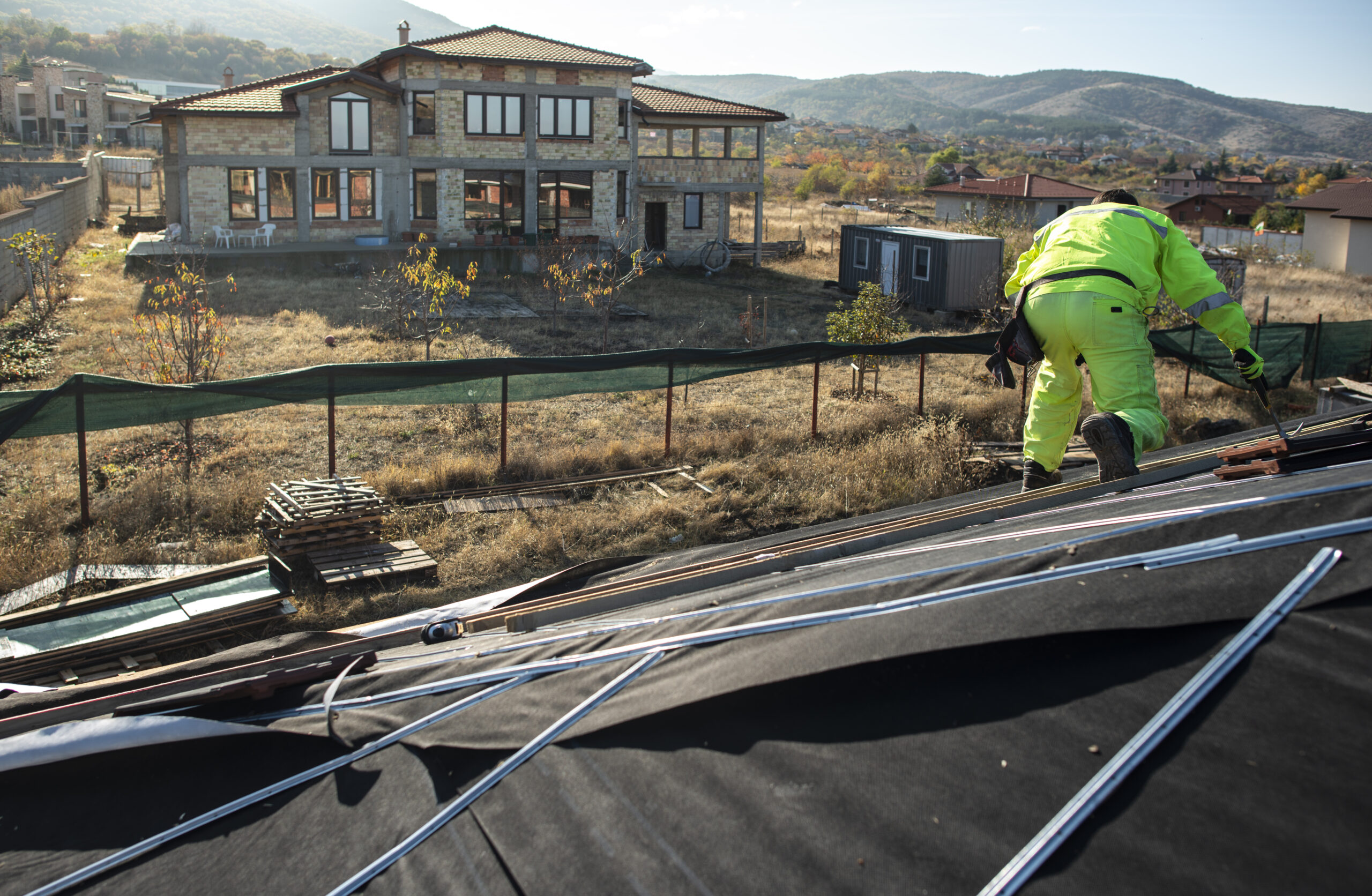 Worker On The Top Of The House Making Roof. Building A New House With Black Roof. Repair And Build Construction Of Roof. Conception For Roofing, New Home, Real Estate And Constructions.